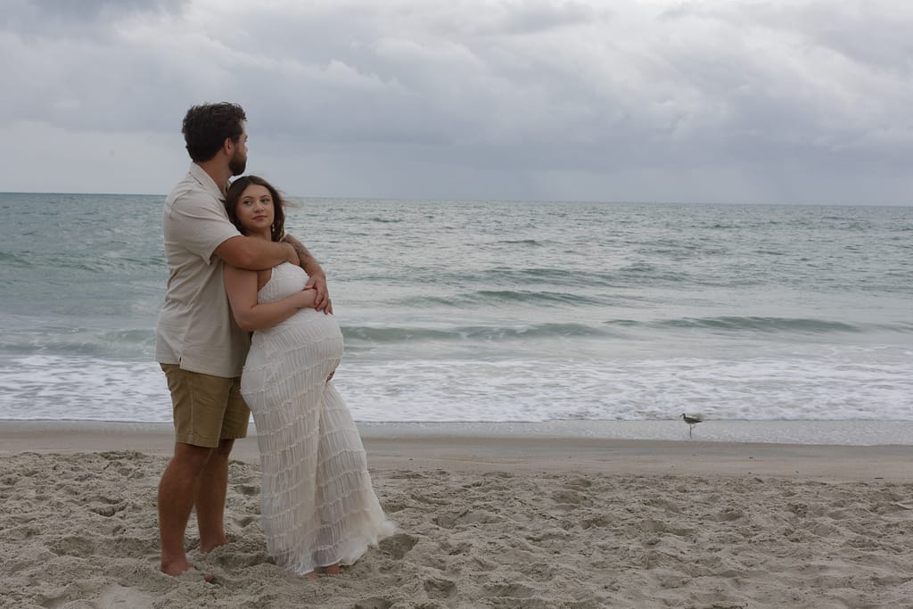 Couple embracing on the shoreline with gray skies above, the sea stretching wide behind them.