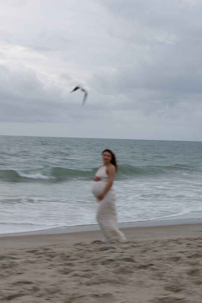 Expecting mother standing on Emerald Isle beach beneath dark storm clouds, wind sweeping her hair as waves crash nearby.